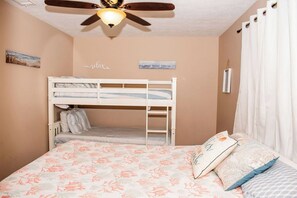 Une chambre avec un lit superposé, un ventilateur de plafond, un lit avec une literie à motifs floraux et un mur orné d’un tableau.