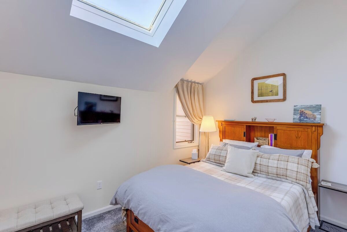 Un dormitorio con cama, un banco, televisor, ventana con cortinas, lámpara y cuadros en la pared.