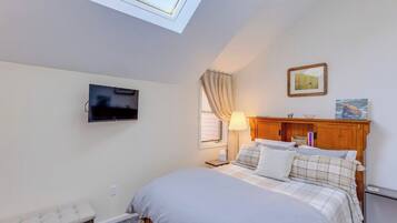 Een slaapkamer met een bed, een bankje, een televisie, een raam met gordijnen, een lamp en kunst aan de muur.