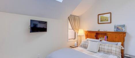 Un dormitorio con cama, un banco, televisor, ventana con cortinas, lámpara y cuadros en la pared.
