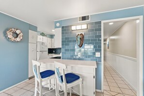 Uma cozinha com armários brancos, painel de fundo azul e um escudo decorativo. Há duas cadeiras brancas com almofadas azuis.
