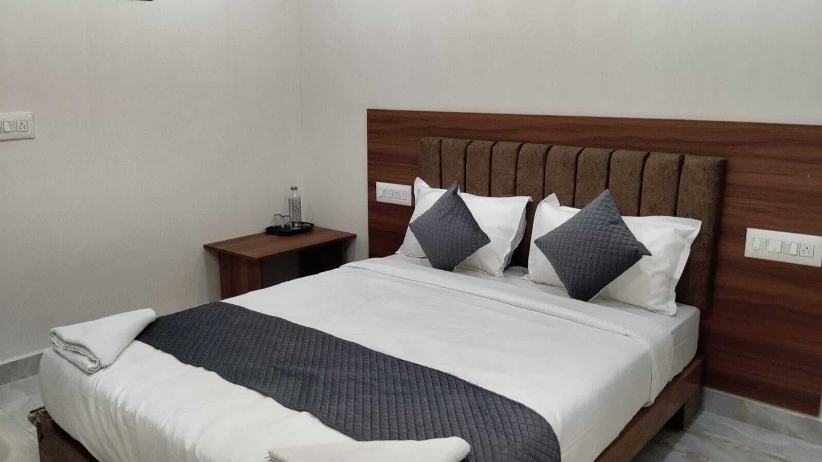 A neatly made bed with a wooden headboard, a bedside table with a bottle and glasses, and a power outlet on the wall.
