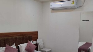 A hotel room with a bed, a nightstand, a chair, an air conditioning unit, and a ceiling fan. - Hotel Udupi Palace (Mysore)
