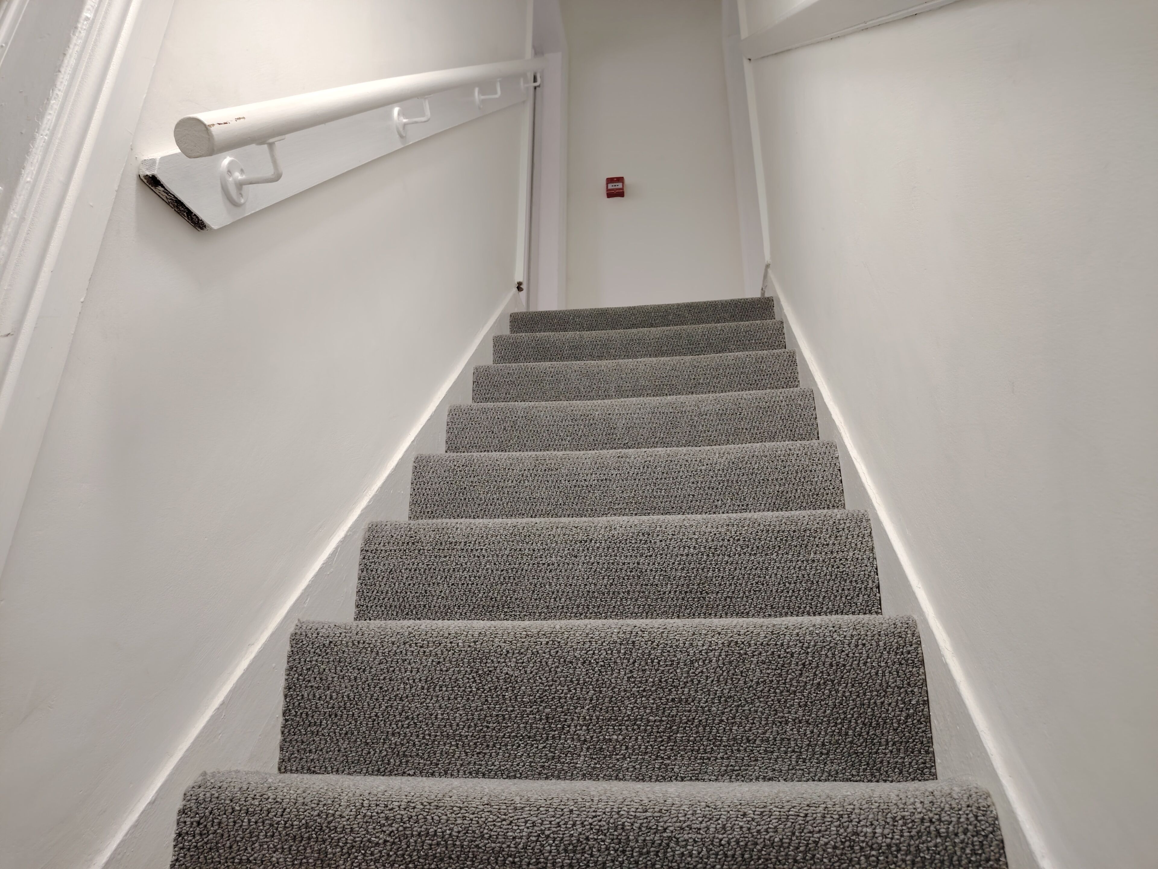 Eine Treppe mit Teppichstufen und einer Handlauf, die zu einer Tür mit Rauchmelder führt.