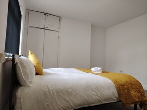 Ein Einzelbett mit weißer Bettwäsche und einer gelben Tagesdecke, ein schwarzer Vorhang und eine weiße Wand mit einer Tür.