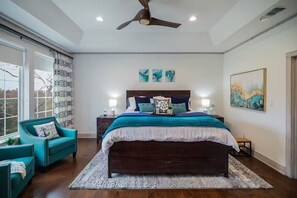 Une chambre avec un grand lit, un ventilateur de plafond, une fenêtre avec des rideaux et des œuvres d’art accrochées aux murs.