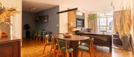 A modern kitchen with a dining area, wooden flooring, and a large window with a city view.