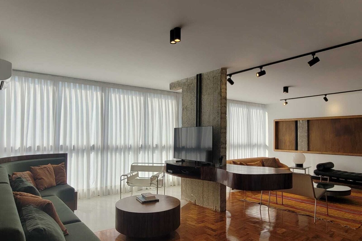 Ein modernes Wohnzimmer mit einer Couch, einem runden Couchtisch, einem Fernseher und einem großen Fenster mit transparenten Vorhängen.