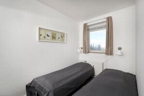 A room with two single beds, a window with curtains, a wall-mounted light, and a framed picture of birds.