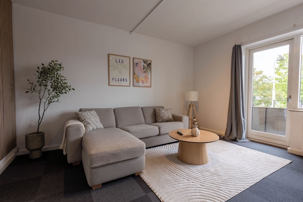 A modern living room with a grey sofa, a small ottoman, a round coffee table, and a potted plant. There are two framed artworks on the wall and a large window with curtains.