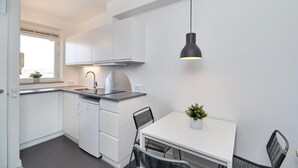 Cocina compacta con armarios blancos, lavavajillas integrado, una mesa de comedor blanca con dos sillas y una lámpara colgante.