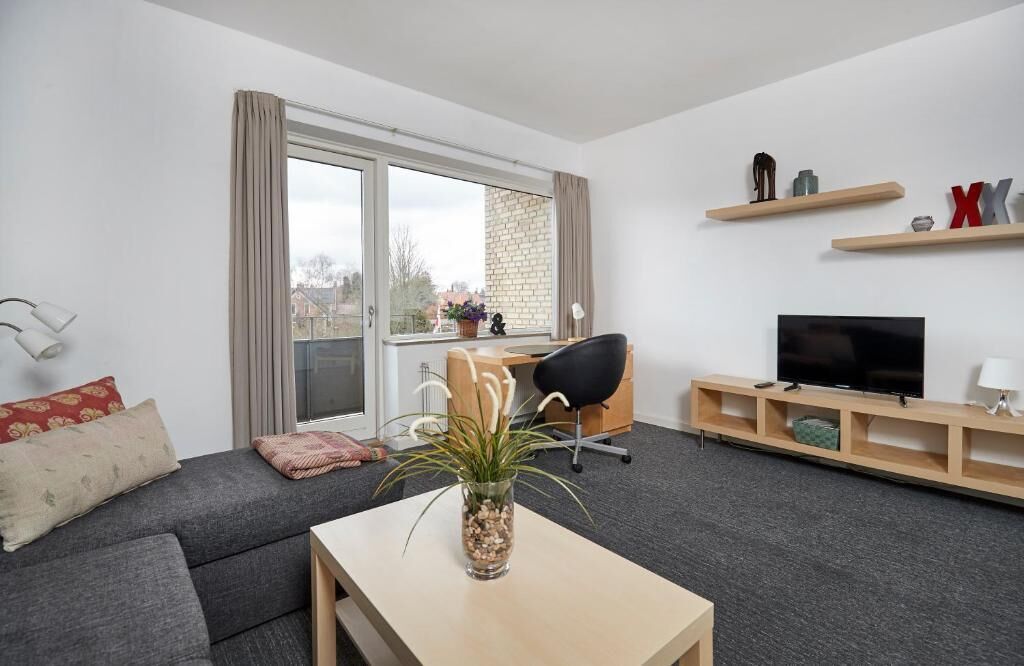 En moderne stue med sofa, sofabord, fjernsyn og en balkon med udsigt til bygninger.
