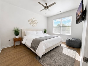 A bedroom with a bed, a ceiling fan, a window with blinds, a wall-mounted TV, and a plant. - Luxury 2BR Near Uptown | Pool, Gym, BBQ (Charlotte)