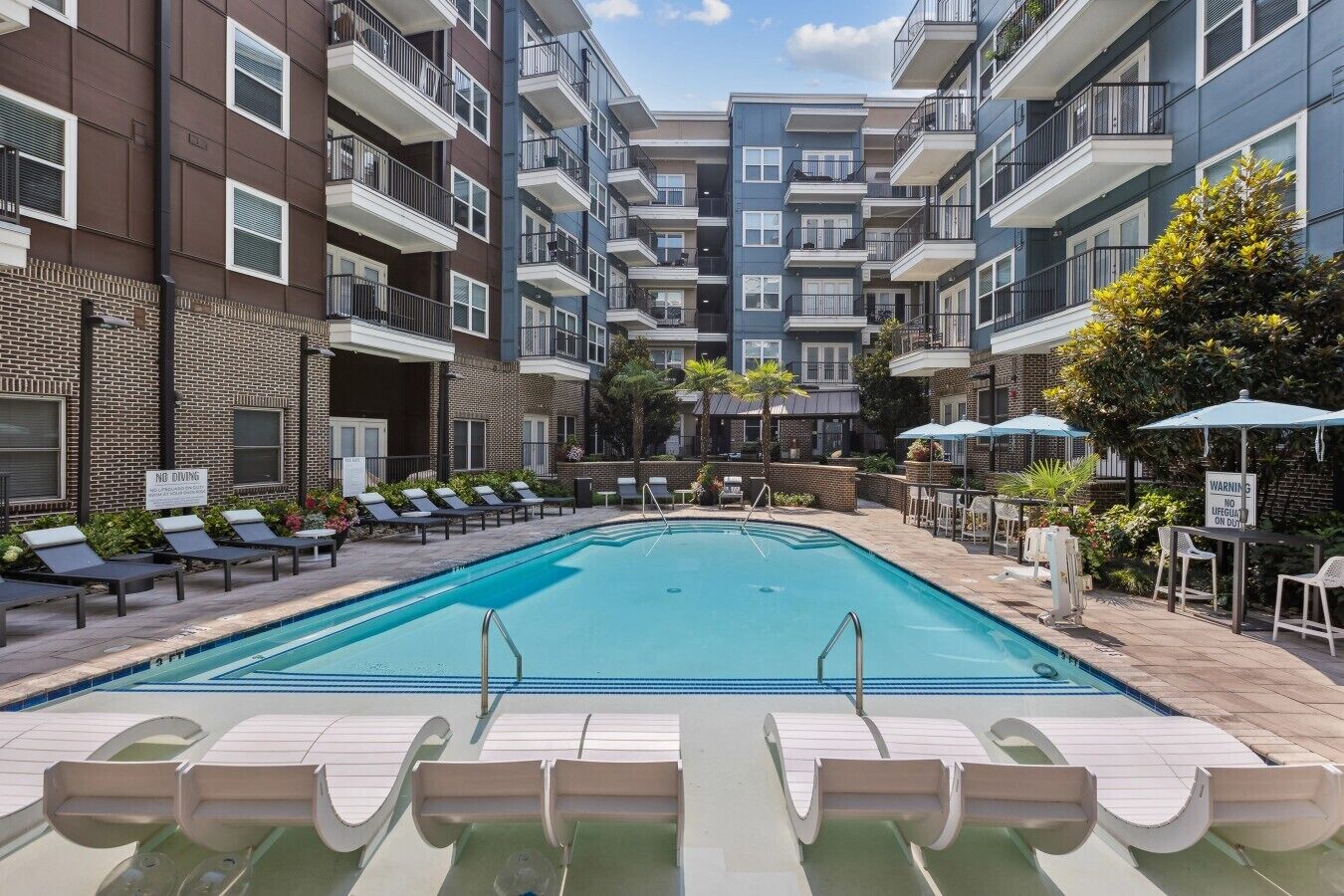A swimming pool with lounge chairs and umbrellas, surrounded by multi-story buildings with balconies.