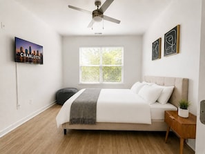 A bedroom with a bed, a ceiling fan, a window with blinds, a TV on the wall, and artwork. - Luxury 2BR Near Uptown | Pool, Gym, BBQ (Charlotte)