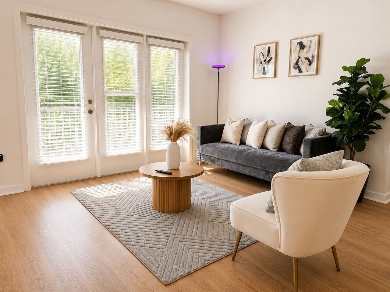 Ein modernes Wohnzimmer mit einer Couch, einem runden Couchtisch und einem Sessel. Zwei Fenster mit Jalousien, eine Stehlampe und eine große Pflanze sind vorhanden.