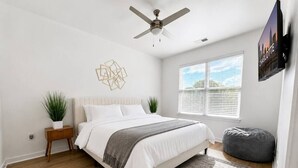 Ein Schlafzimmer mit einem Bett, einem Deckenventilator, einem Fenster mit Jalousien, einem an der Wand montierten Fernseher und einer Pflanze.