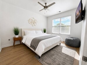 Quarto com cama, ventilador de teto, janela com persianas, televisão na parede e uma planta.