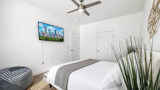 Ein modernes Schlafzimmer mit einem Bett, einem Deckenventilator, einem Stadtbild-Fernseher und einer Pflanze.