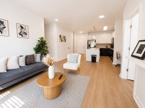 Ein modernes Wohnzimmer mit einer Couch, einem Couchtisch und einer Pflanze. Im Hintergrund befindet sich eine Küche mit Schränken und einem Kühlschrank.