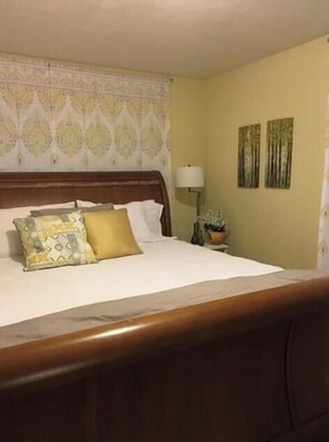 Habitación de hotel con cabecera de madera, cama con ropa blanca y almohadas estampadas, lámpara de noche y dos cuadros colgados en la pared.