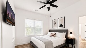 Een slaapkamer met een bed, een plafondventilator, een nachtkastje, een tv aan de muur en een raam met zonwering.