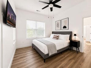 Ein Schlafzimmer mit Bett, Deckenventilator, Nachttisch, Wand-Fernseher und Fenster mit Jalousien.