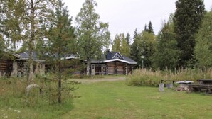Hirsimökki, jossa on kuisti, ja sen ympärillä on nurmikkoalue ja puita.