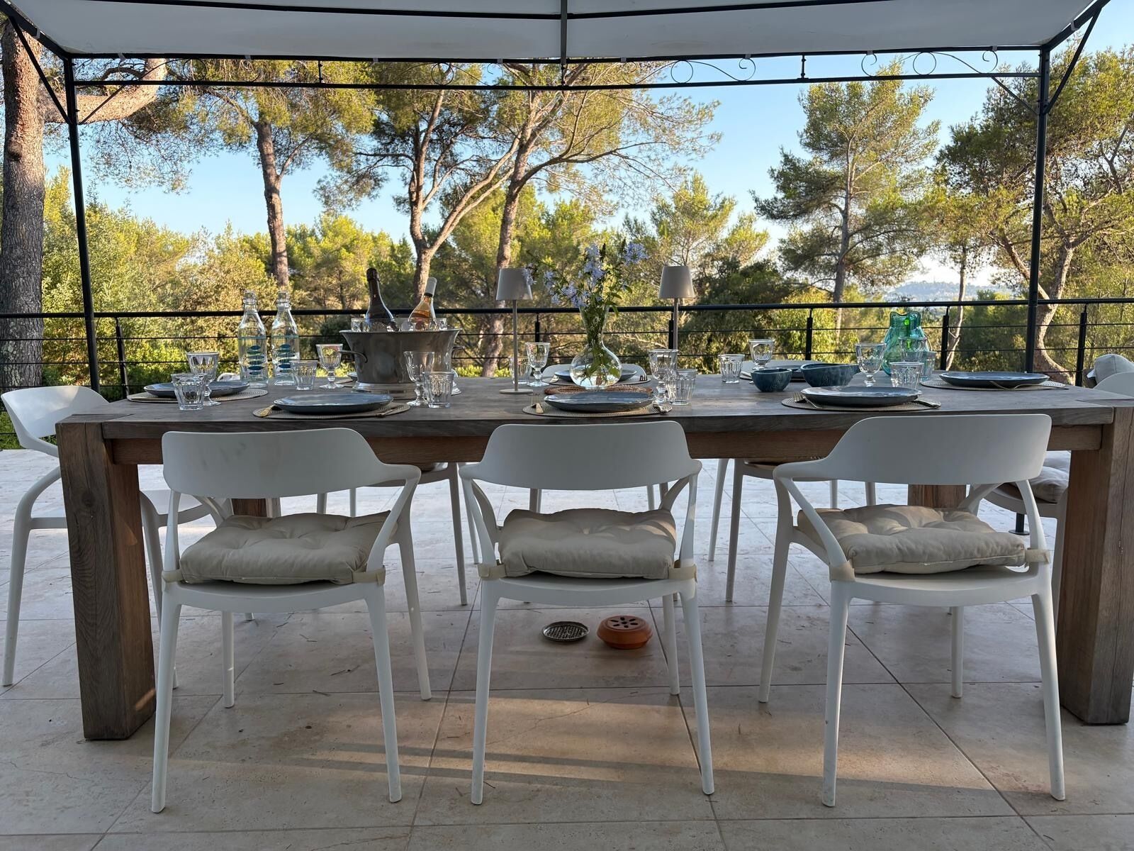 An outdoor dining area with a table set for four, surrounded by white chairs with cushions, under a covered structure with a view of trees.