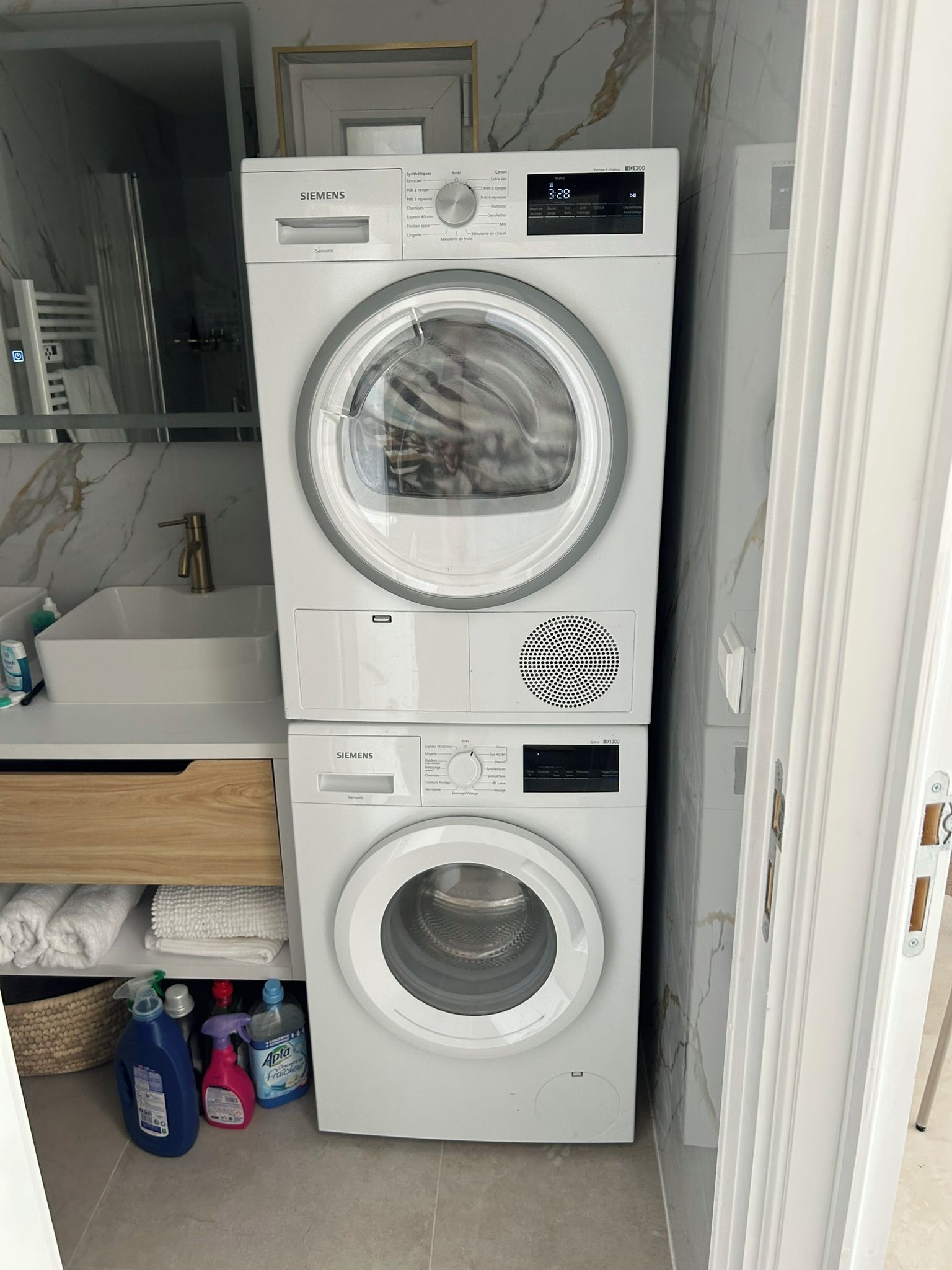 Une machine à laver et un sèche-linge superposés dans une salle de bain, avec un pare-éclaboussures en marbre et un lavabo où sont posés des articles de toilette.