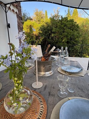 An outdoor dining setup with a table, metal bucket, wine bottles, glasses, and a decorative vase with flowers. - Villa Orange (La Croix-Valmer)