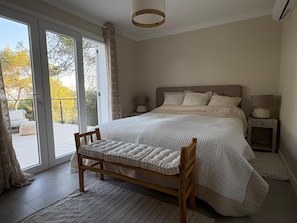 A bedroom with a large bed, a bench, a nightstand, and a view through a sliding glass door. - Villa Orange (La Croix-Valmer)