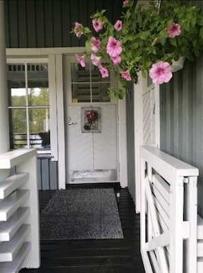Eine Veranda mit einer weißen Tür, einer schwarzen Fußmatte und einem hängenden Blumenarrangement.