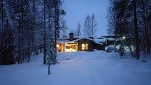 A snow-covered landscape with a cabin and a car parked in the driveway. - Höperö | Charming 1BR Apt | Sauna & Fireplace (Ruka)
