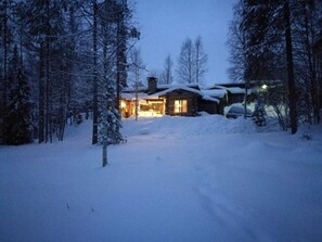A snow-covered landscape with a cabin and a car parked in the driveway. - Höperö | Charming 1BR Apt | Sauna & Fireplace (Ruka)