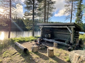 A rustic cabin with a fire pit and logs, surrounded by a forest and a body of water. - Mestarikämpät B 9 | Cozy Lakeside Apt with Sauna (Ruka)