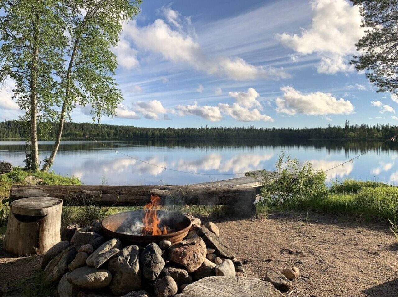 A campfire with rocks and logs, a wooden log, and a calm lake with trees in the background.