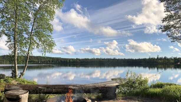 Nuotiopaikka kivien ja puiden ympäröimänä, tukki ja tyyni järvi, jonka taustalla näkyy puita.