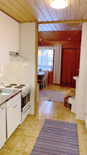 A compact kitchen with white cabinets, a stove, and a dining area with a table and chairs. - Mestarikämpät B 9 | Cozy Lakeside Cottage & Sauna (Ruka)
