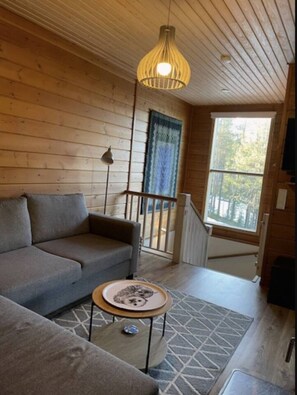 Ein Holzinnenraum mit einer Couch, einem kleinen runden Tisch, einer Hängelampe und einem Fenster mit Blick auf Bäume.