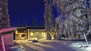 Lumipeitteinen maisema, jossa on rakennus, jossa on suuri ikkuna ja ovi, ja rakennuksen ympärillä on puita.