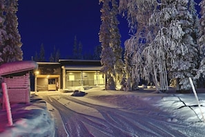 A snow-covered landscape with a building featuring a large window and a door, surrounded by trees. - Villa Viihy | Modern 2BR Villa Near Ski Slopes (Ruka)