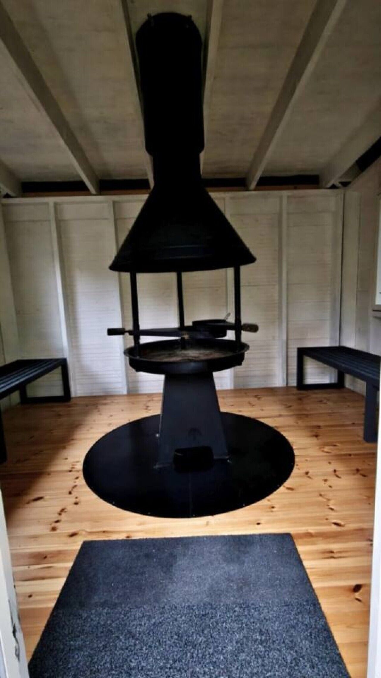 Een kamer met een centrale, zwarte kachel, houten vloeren en een zwarte mat bij de ingang.