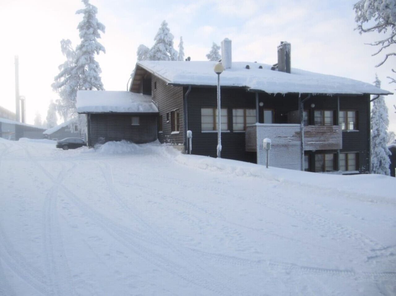 En snötäckt trästuga med en synlig skorsten, omgiven av snötyngda träd.