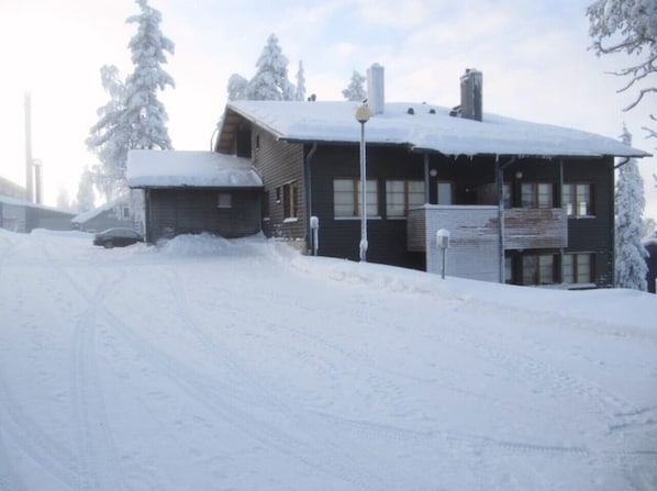 Une cabane en bois recouverte de neige, avec une cheminée bien visible, entourée d’arbres chargés de neige.
