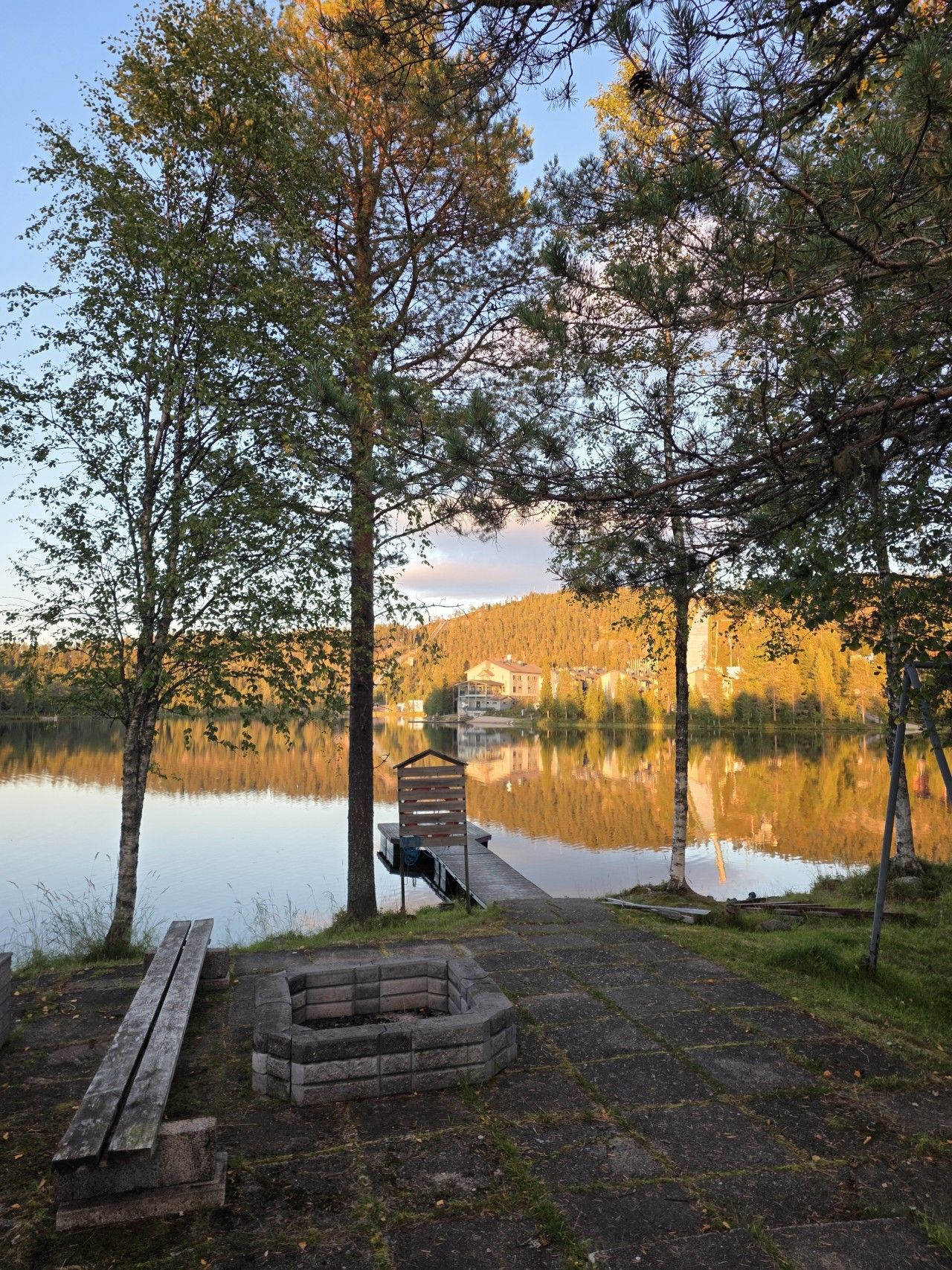 Una cabaña junto al lago con muelle, una fogata y un banco.