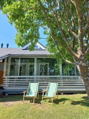 Ein Haus mit überdachter Veranda, zwei gestreiften Liegestühlen und einem Baum im Vordergrund.