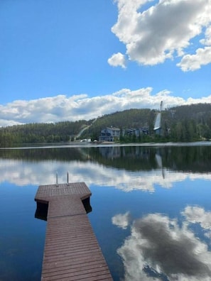 Ein ruhiger See mit einer hölzernen Anlegestelle, die sich ins Wasser erstreckt, umgeben von einem bewaldeten Ufer und einem Gebäude mit geneigtem Dach.