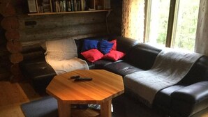 Een gezellige woonkamer met een houten salontafel, een zwarte leren bank met rode en blauwe kussens, en een boekenkast tegen de muur.