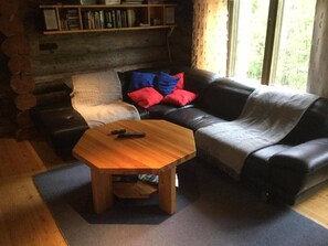 A cozy living room with a wooden coffee table, a black leather sofa with red and blue cushions, and a bookshelf against the wall. - RukanTykky | Charming 3BR Chalet | Near Slopes (Ruka)